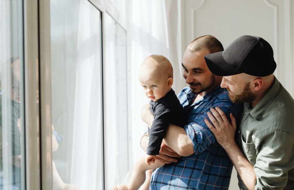 Baby and 2 dads smiling next to a window