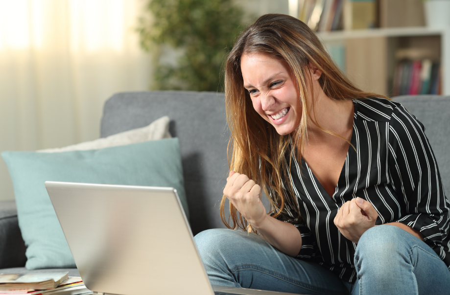 A woman with long hair, a stripy top and jeans, looking like she has had a victory and is celebrating at something she is looking at on her laptop, which is on a coffee table in front of her